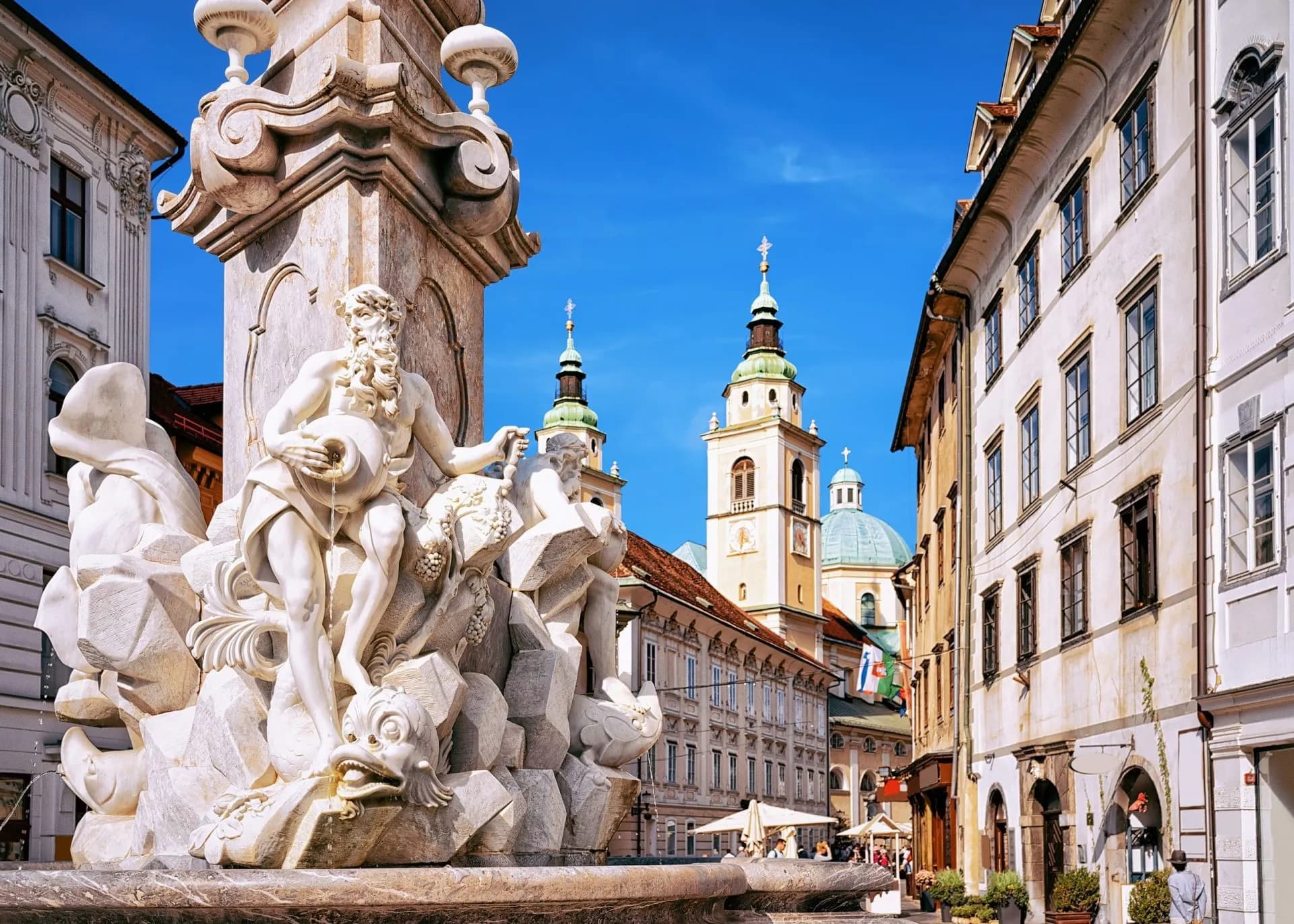 Baroque fountain sculpture in Ljubljana square with church towers under a bright blue sky.